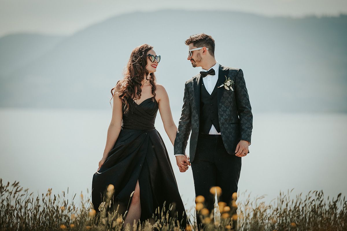 A bride in a black wedding gown holds her groom's hand as they walk in a field of tall grass. The groom wears a black suit and bow-tie.