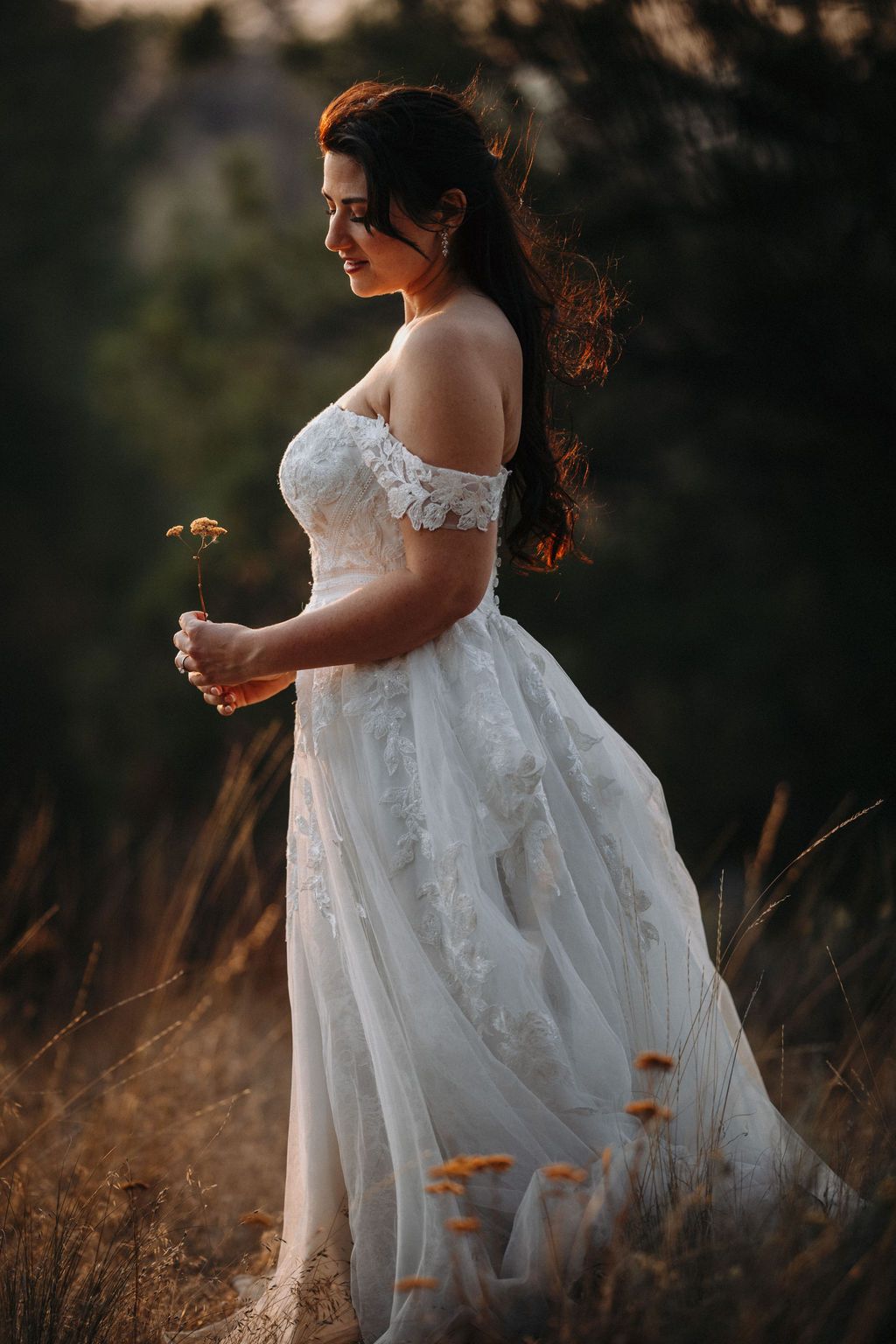 A bride wearing a flowing white wedding dress moves gracefully through a field of tall grass at golden hour.