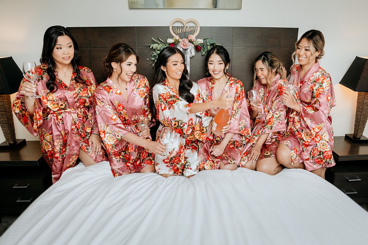 A group wearing matching floral pink robes sits together on a white bed during a pre-wedding celebration.