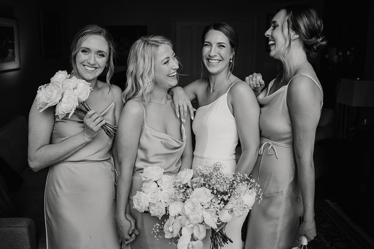 A bridal party shares a candid moment of laughter while holding elegant floral bouquets at a wedding celebration. Black and White.
