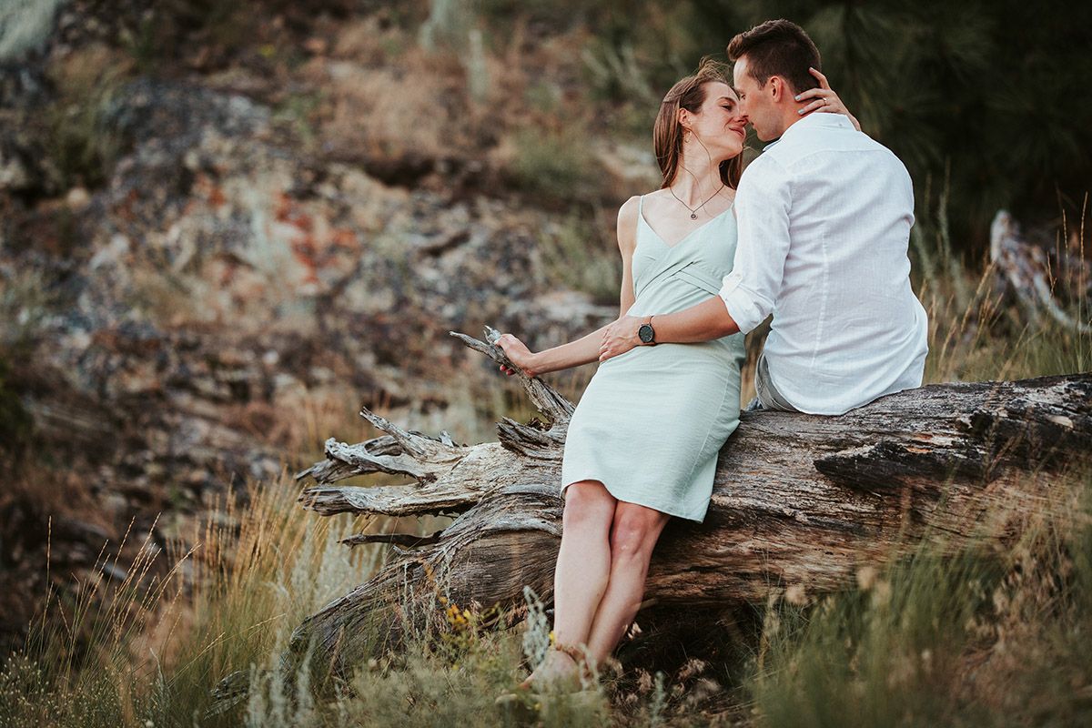 Couple sharing a romantic kiss while sitting on a fallen log in a natural outdoor setting.
