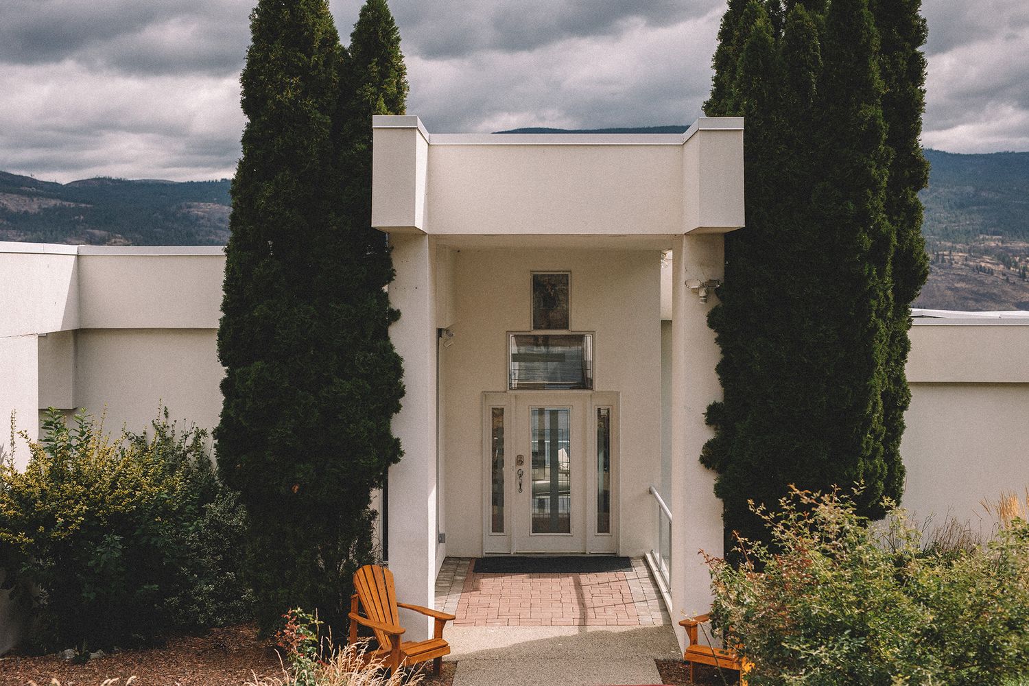 Modern white villa with tall cypress trees against cloudy sky and mountain backdrop in Mediterranean setting.