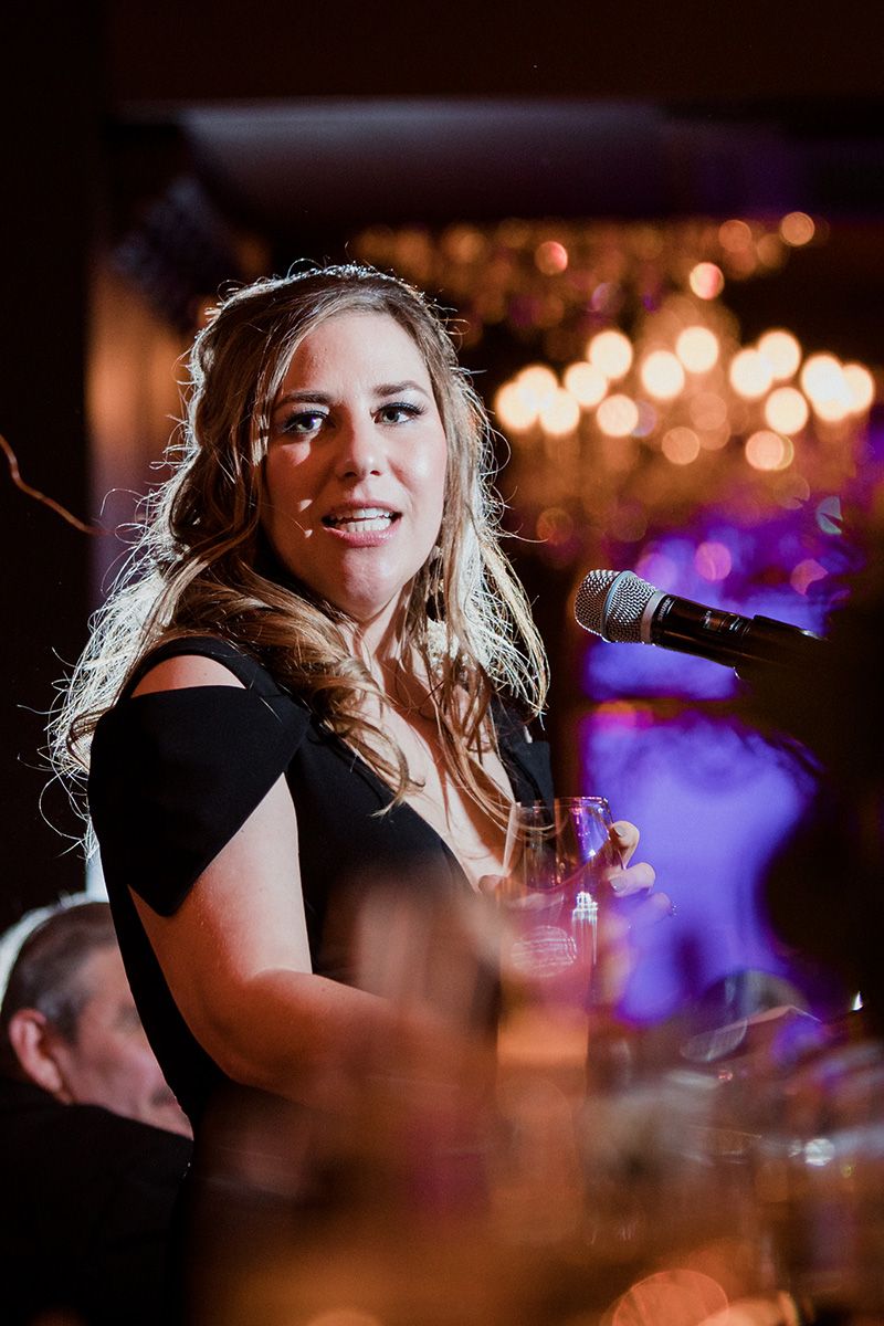 Performer in black top smiling and singing on stage with blurred bokeh lights and purple ambiance in background.