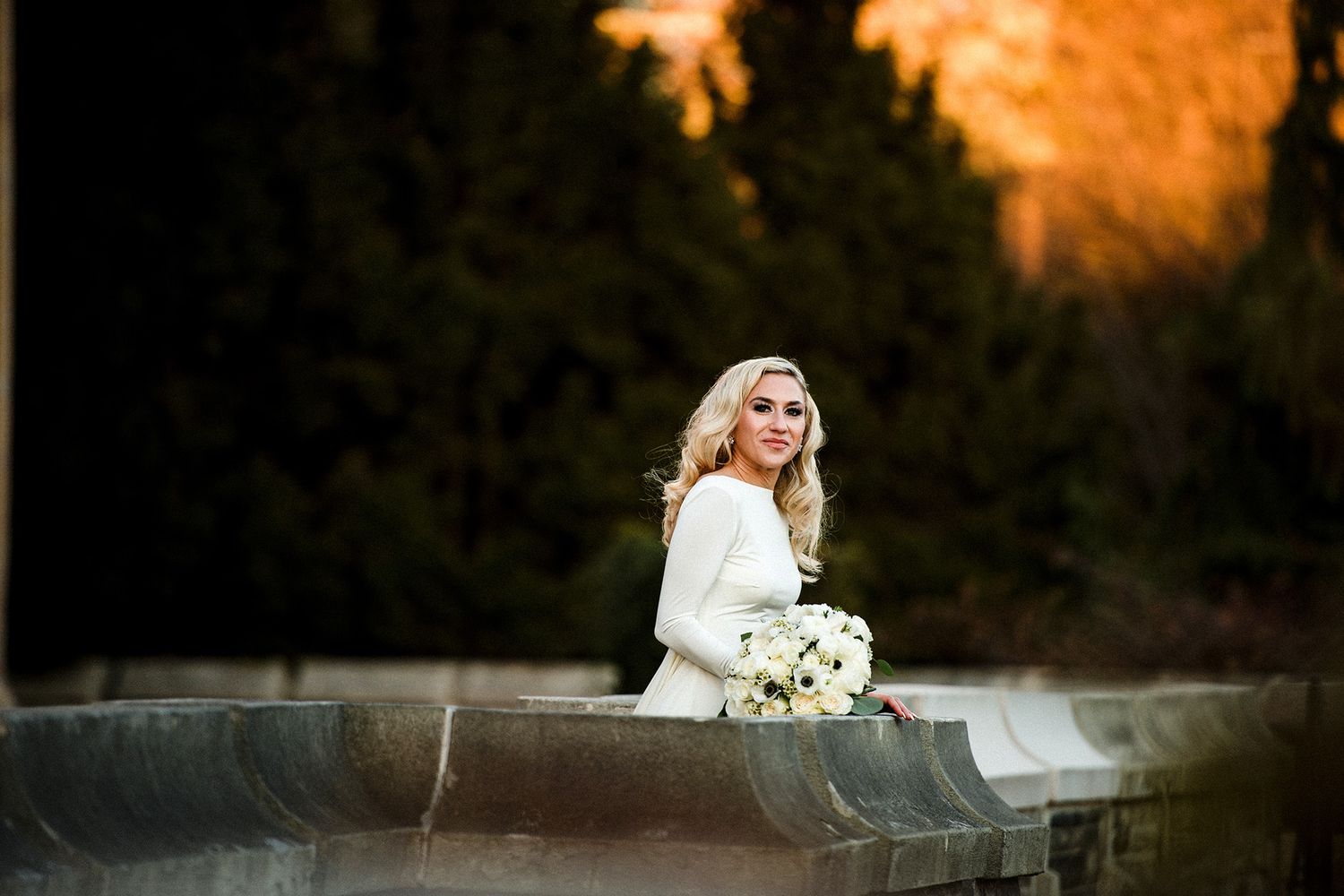 Bride in white dress sits on concrete wall holding white bouquet during golden sunset.