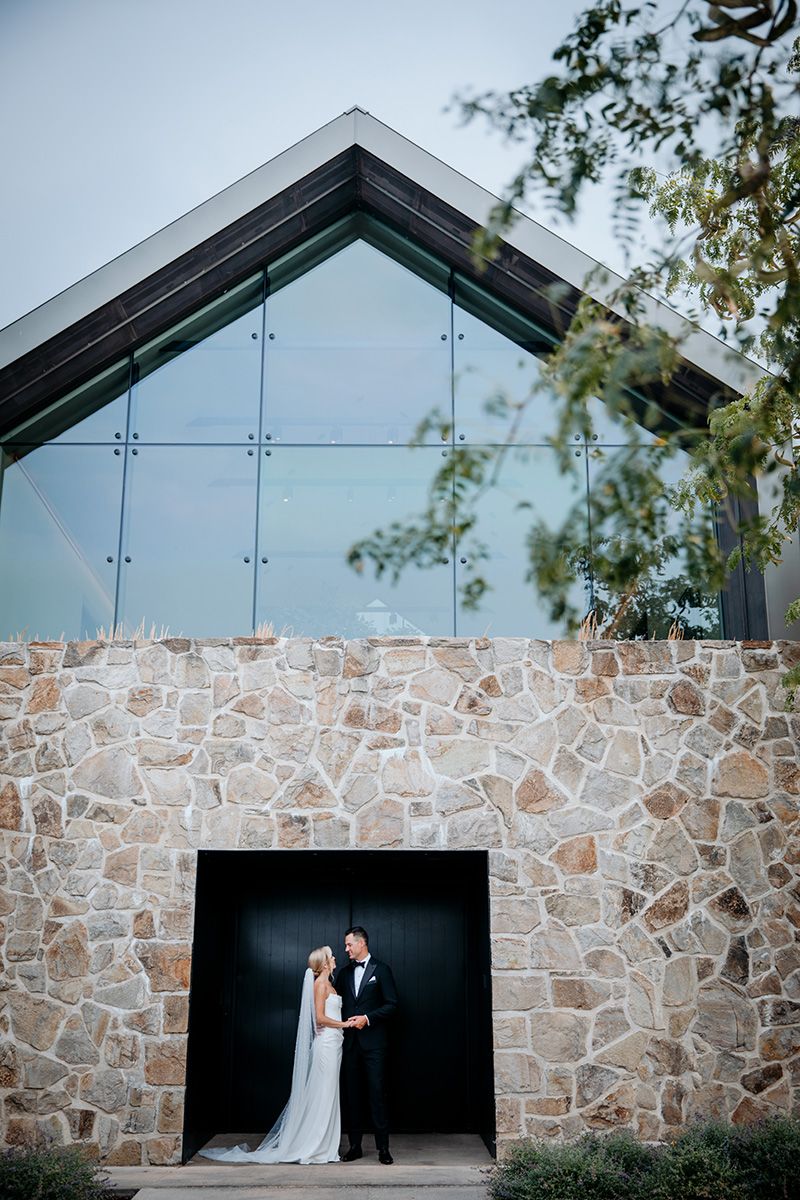 Bride and groom appear small in a long shot of a modern stone and glass chapel with dramatic angular architecture and black doorway.