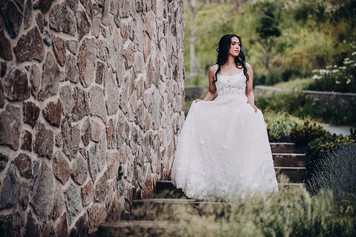 A bride in a white lace gown poses against a rustic stone wall in an outdoor garden setting.