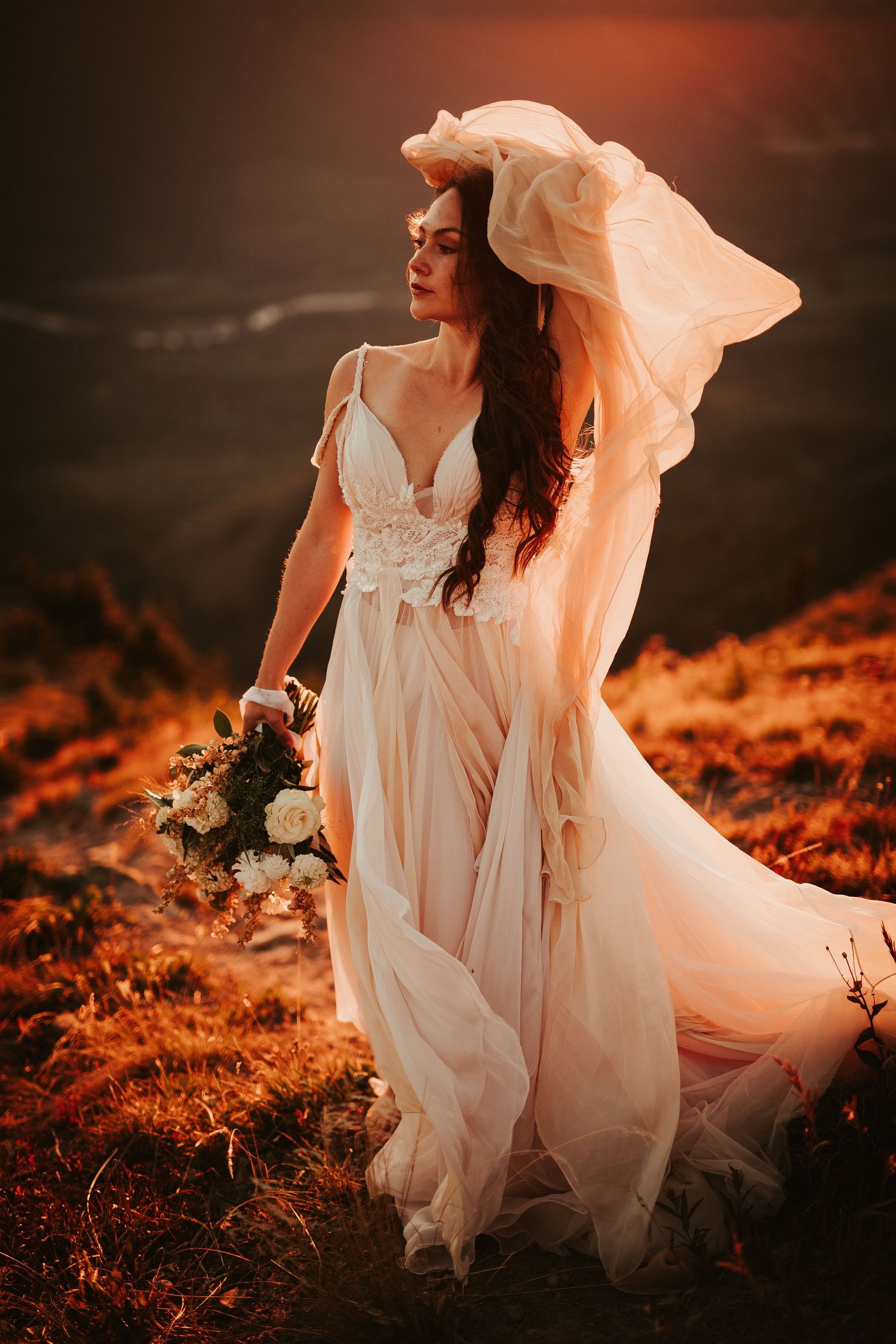 A flowing white wedding dress billows in the wind against a dramatic sunset-lit mountainous landscape.