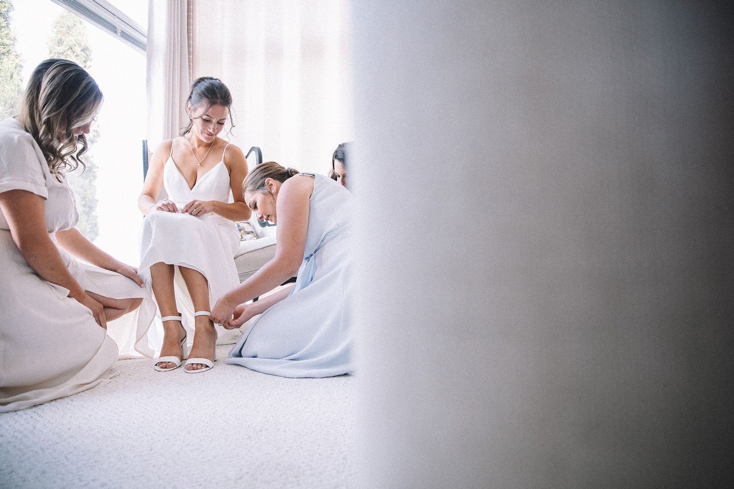 A group of people in white clothing sit together on a bed during a wedding preparation moment.