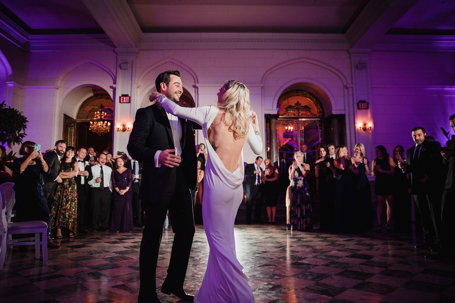 Couple shares their first dance in a purple-lit ballroom with elegant architecture and watching guests.