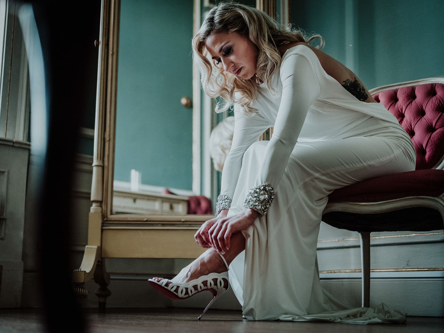 A bride in a white wedding dress sits gracefully on a pink velvet couch as she adjusts the strap of her high-heel shoe.