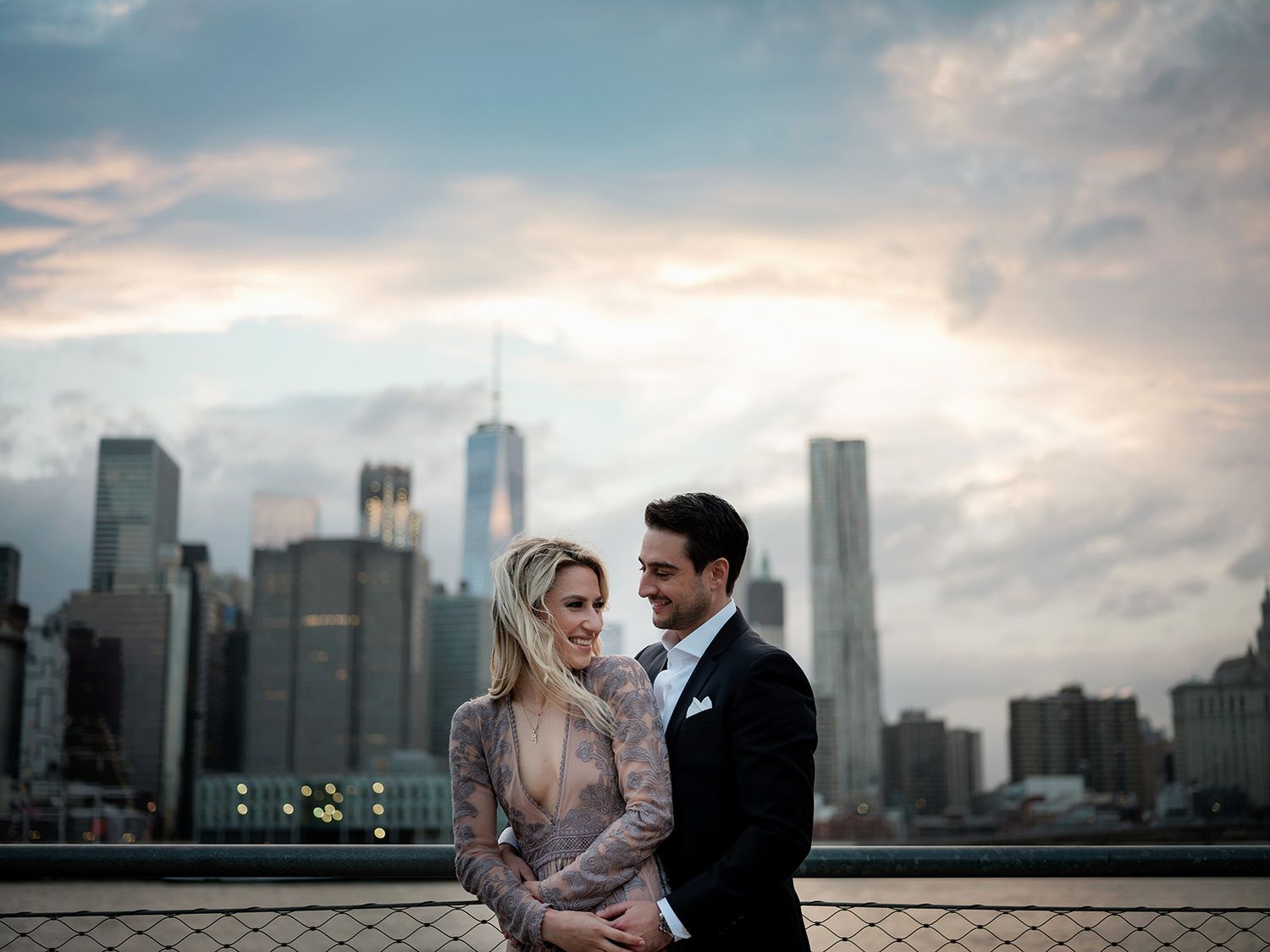 A couple embraces against the backdrop of New York City's skyline at dusk with One World Trade Center visible.