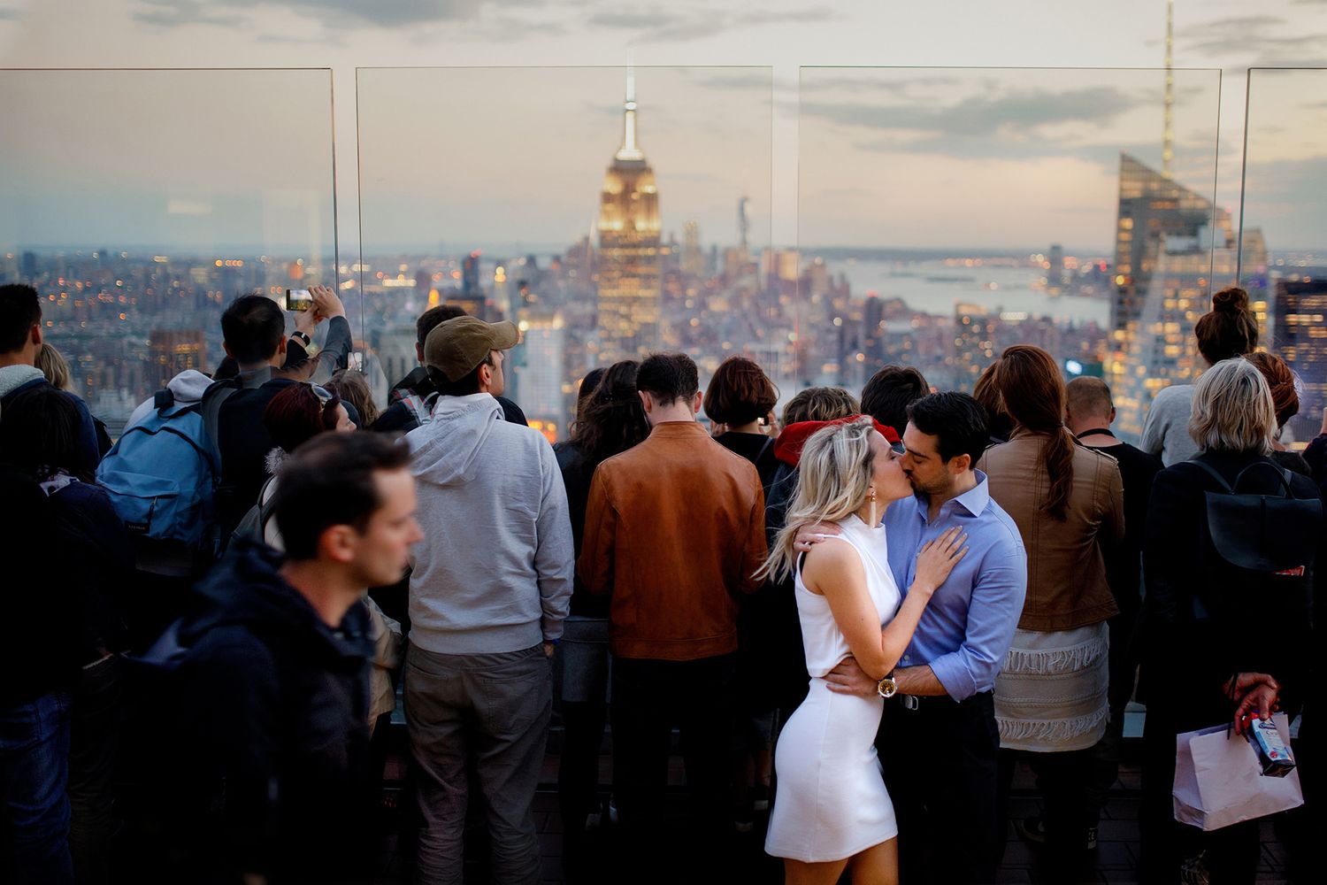 Crowd of people enjoying sunset views from Top of the Rock observation deck overlooking New York City skyline while a couple share a kiss.