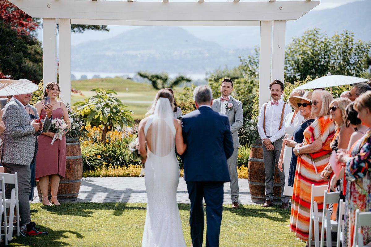 An outdoor wedding ceremony moments showing guests and decorations under a white pergola on a sunny day.