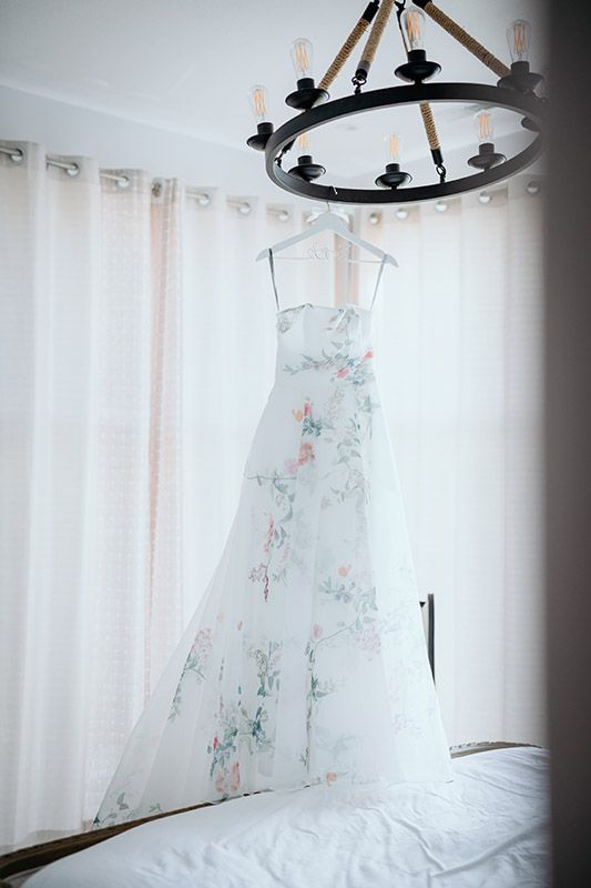 A delicate white floral dress hangs from a rustic chandelier against sheer curtains in a bright room.