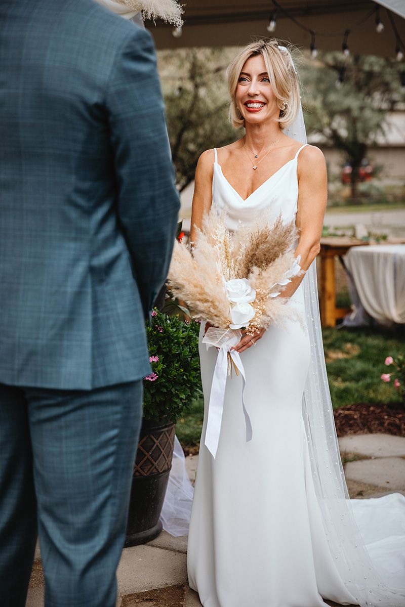 A bride in a white dress and groom in a blue suit during a sunny outdoor ceremony.