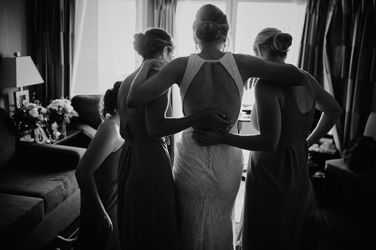 Black and white photo from behind a bride and her maids in a dimly lit room facing the window.