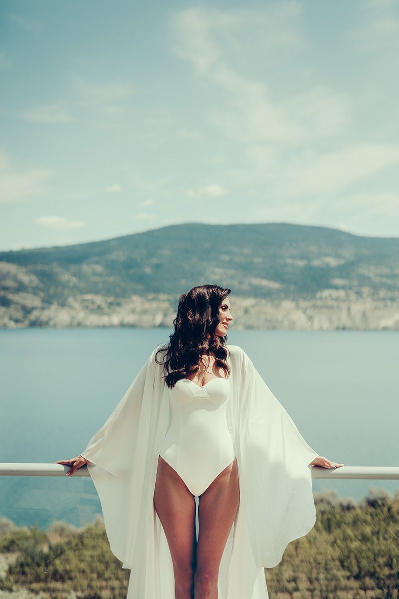 Woman in white flowing cape poses gracefully against scenic coastal mountain landscape at sunset.