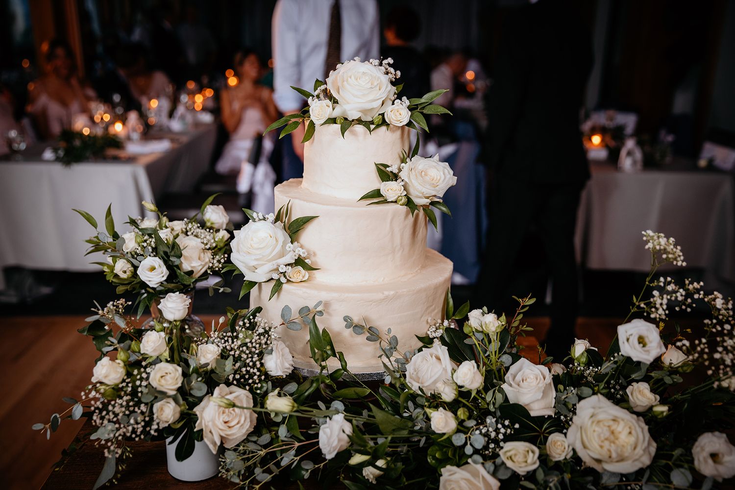 Elegant white wedding cake decorated with fresh roses and greenery sits on display at an evening reception.