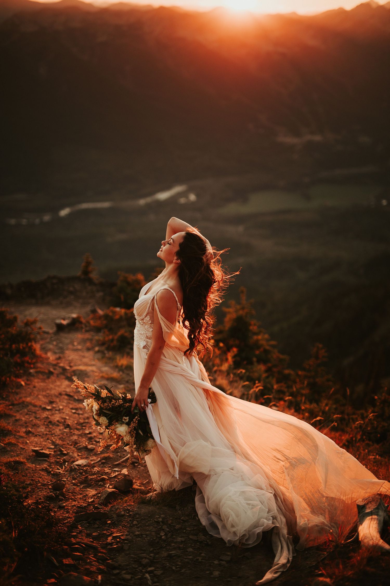 A flowing white wedding dress catches the light at sunset on a mountain overlook as wind sweeps through the scene.