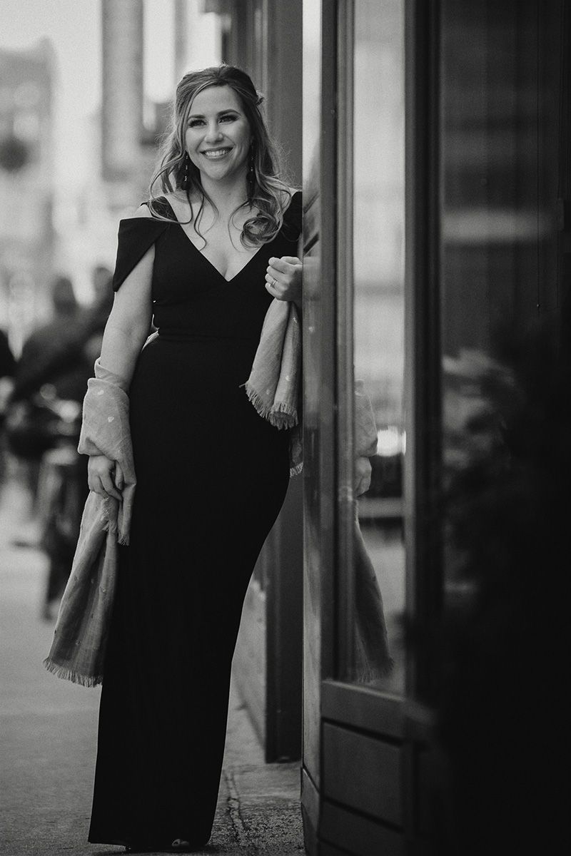 A black and white photos showing a woman in a black dress standing against a building exterior.