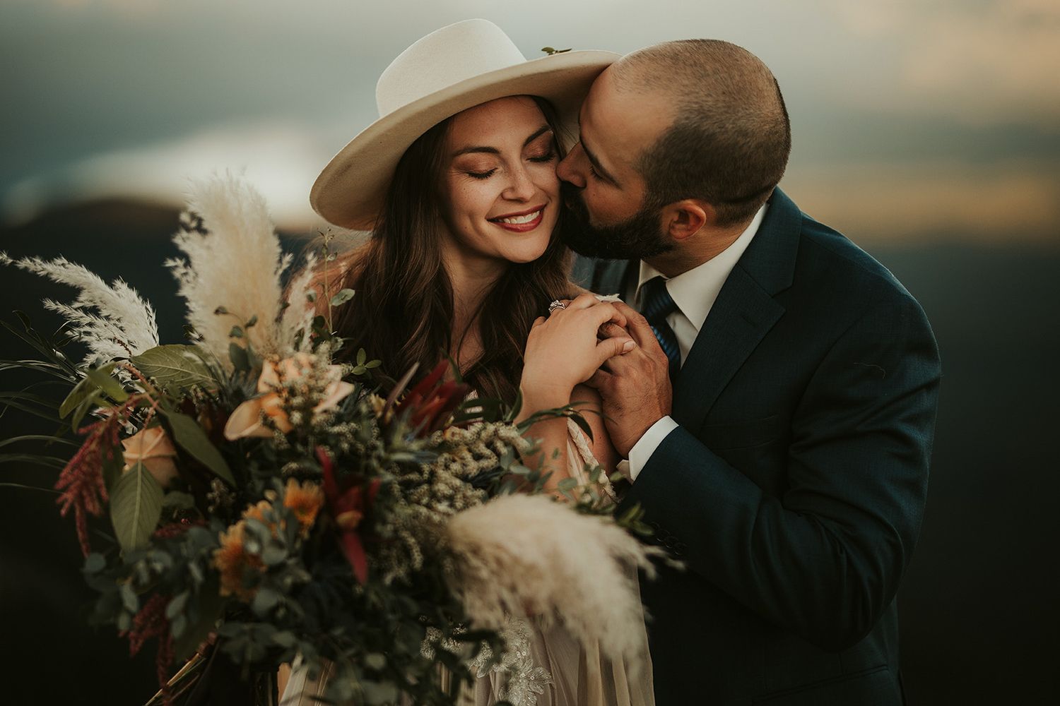 A romantic couple embraces during a sunset wedding photo shoot with dramatic clouds in the background.
