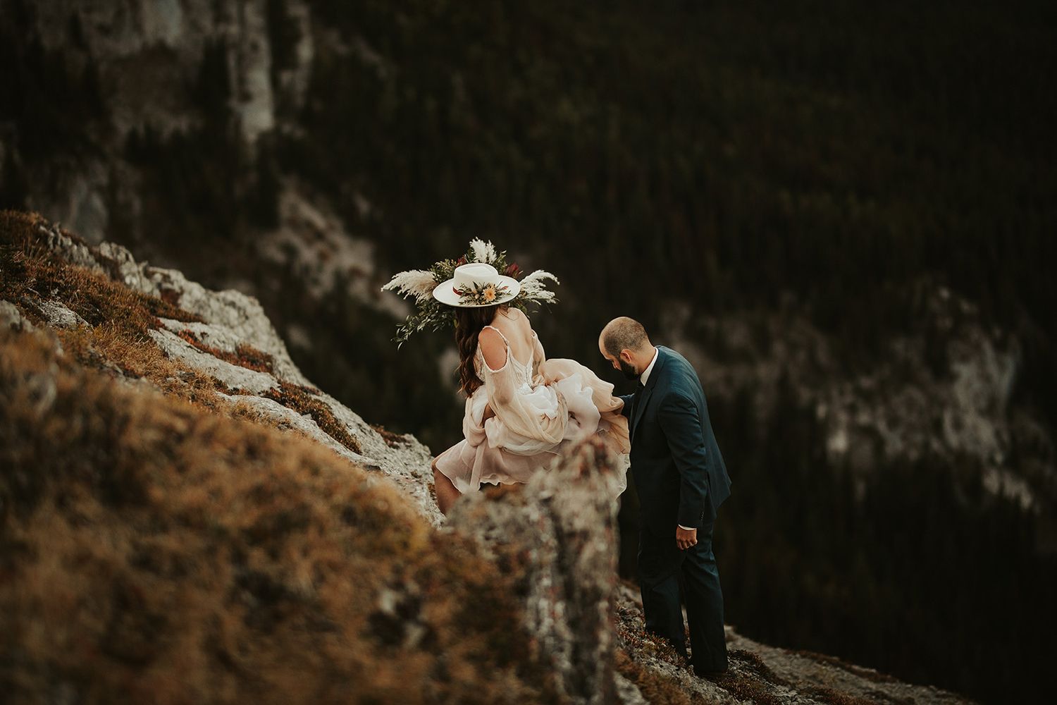 A groom holds his bride's dress as they both negotiate a a cliff edge during a sunset wedding photo shoot.