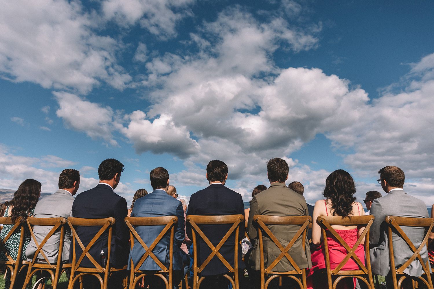 Row of wedding guests seated in wooden chairs watching ceremony against blue sky with white clouds.
