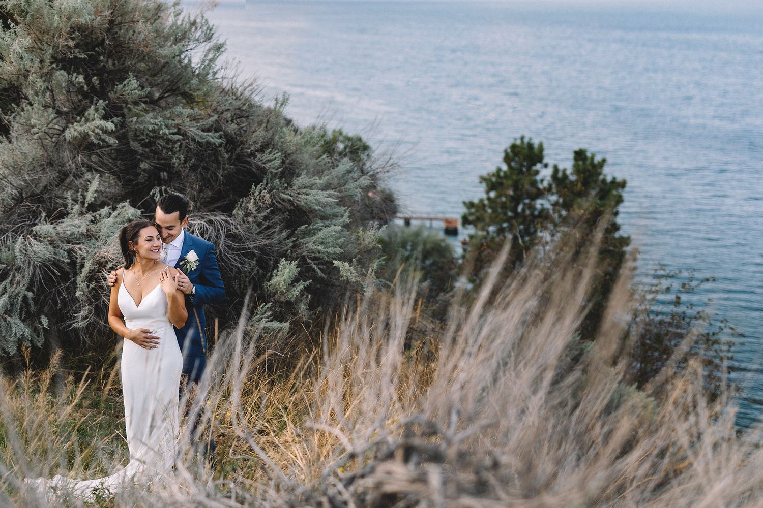 Romantic coastal wedding photoshoot with couple embracing among tall grass and ocean views at sunset.