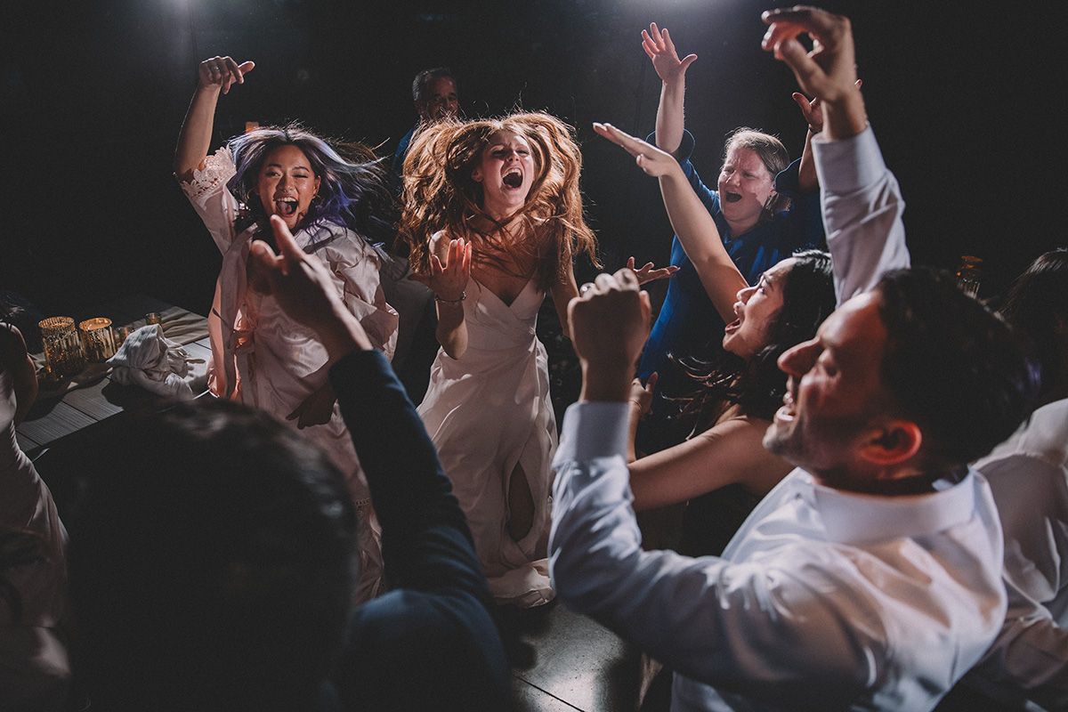 A crowd of people dancing energetically with raised arms at a wedding reception in moody lighting.