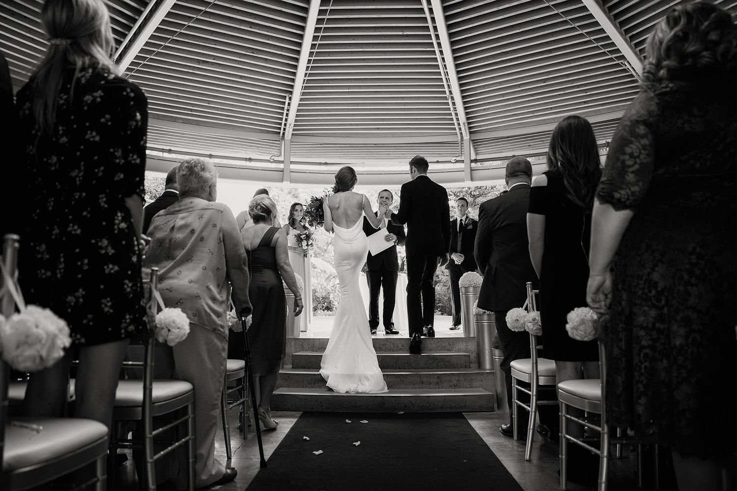 A black and white photo of a wedding ceremony with the bride and groom standing on steps in an architectural space with curved lines.