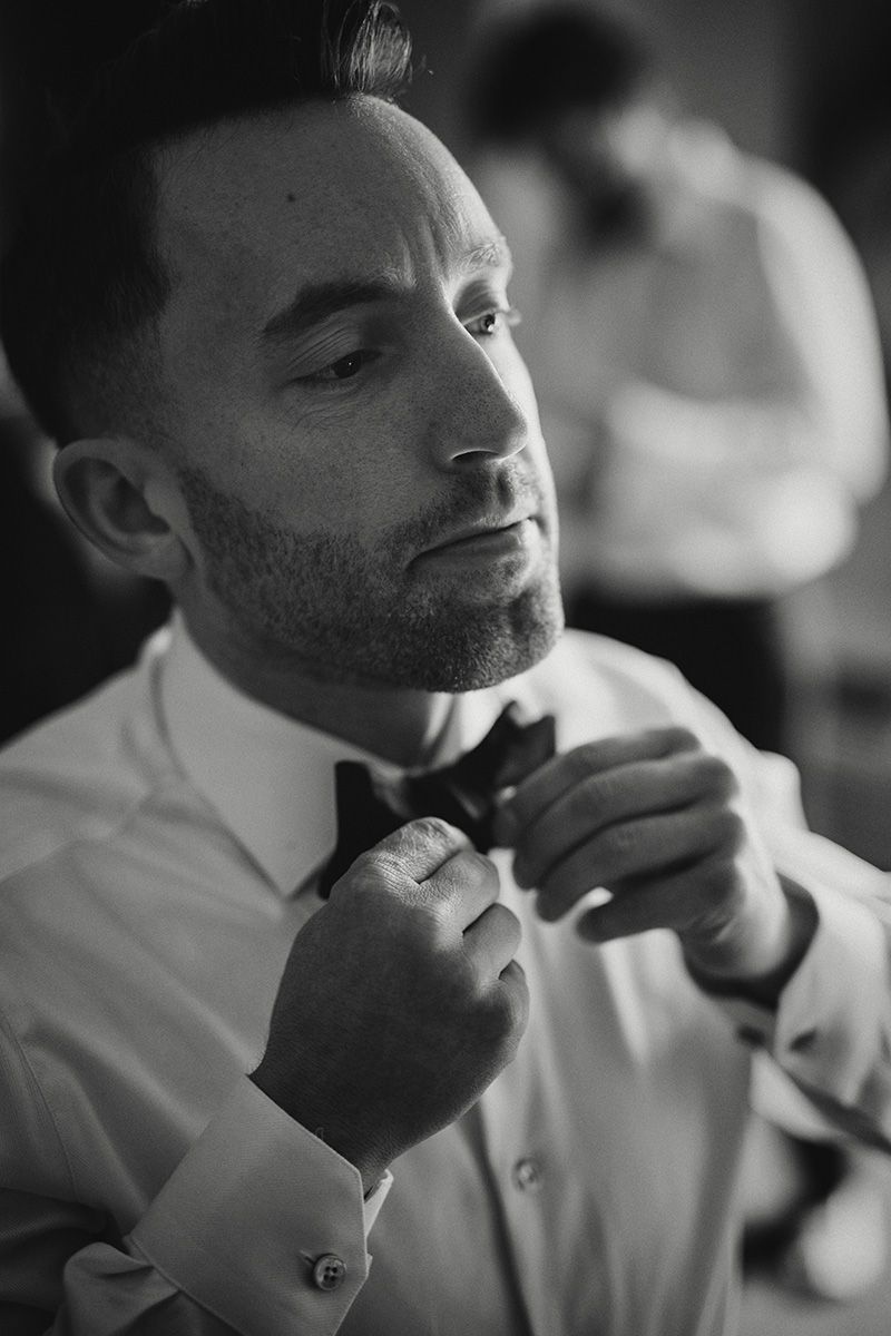 A black and white photo of a groom adjusting his bow-tie while getting dressed in formal attire.