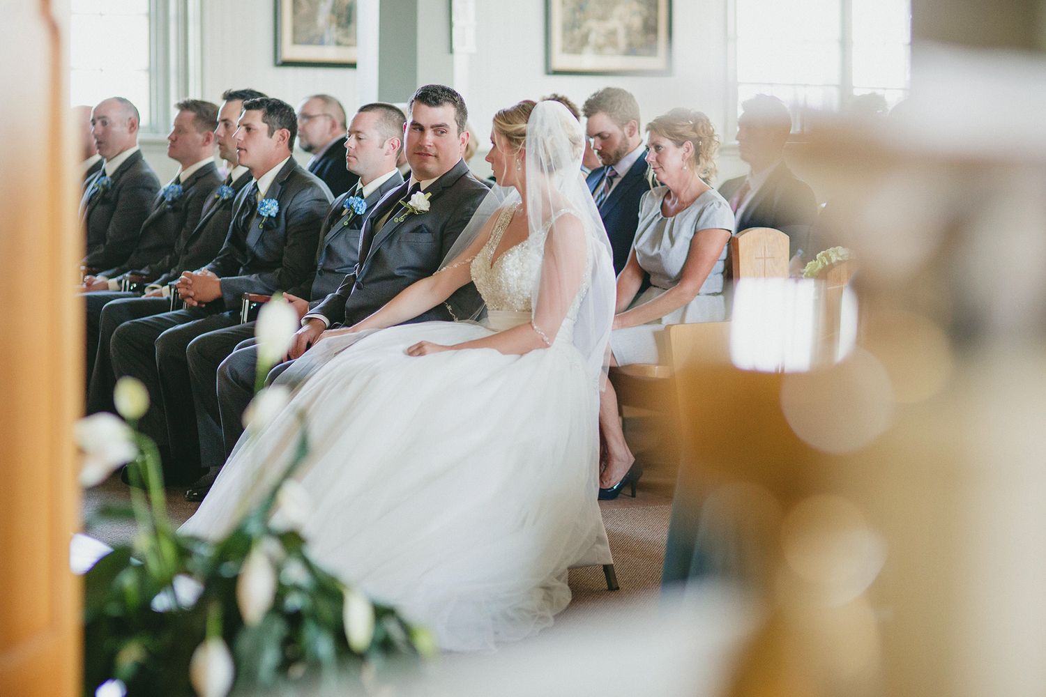 A bride in a white wedding gown sits in the front row during a church ceremony with guests seated behind her.