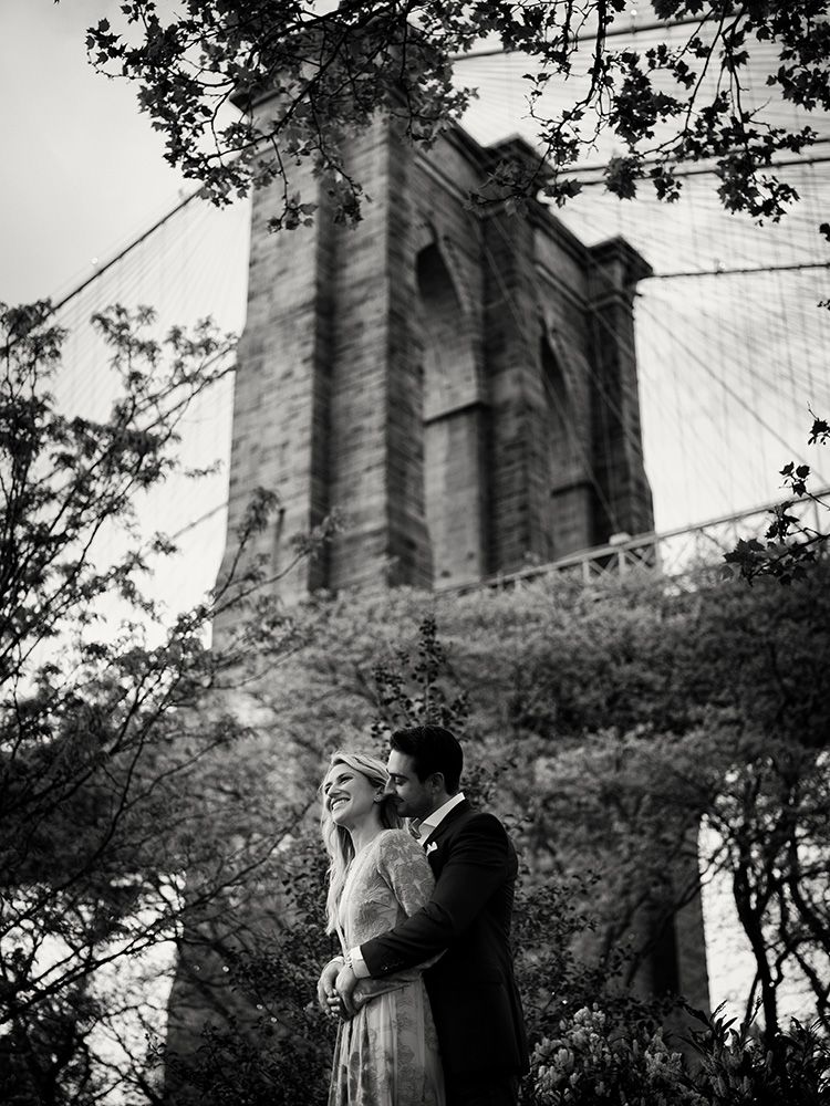 A romantic black and white photo of a couple embracing near the Brooklyn Bridge with dramatic architectural angles.