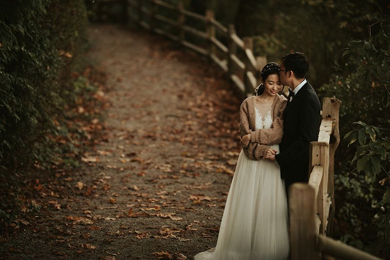 A couple shares an intimate moment on a rustic forest path surrounded by autumn foliage and lantern lighting.