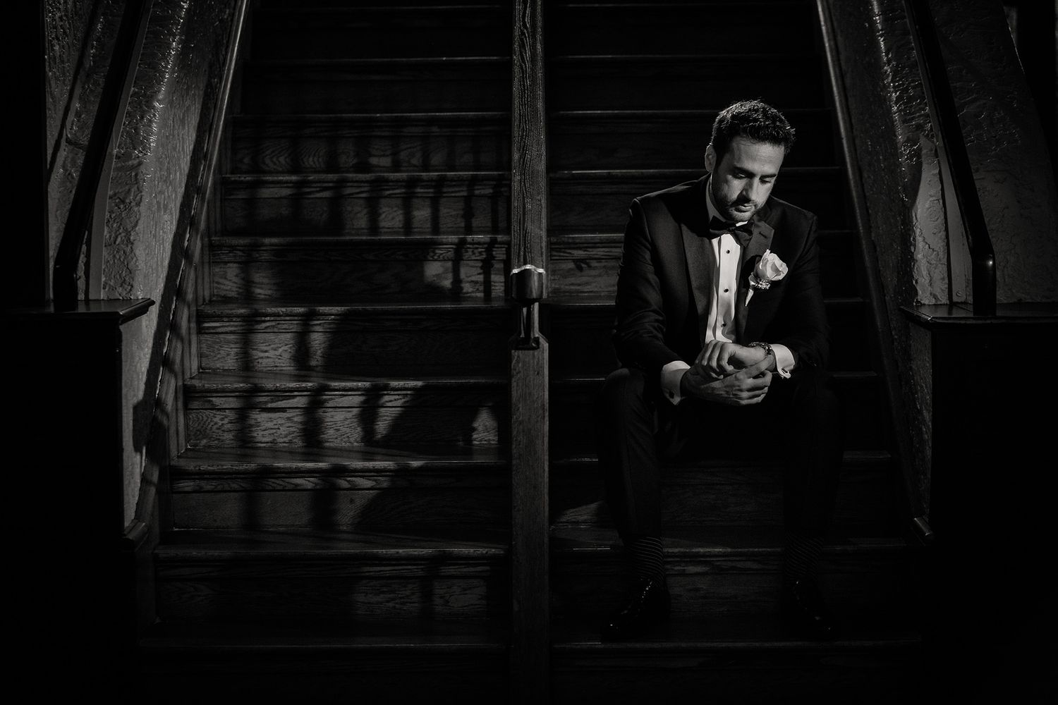 A black and white wedding photo showing a groom in a tuxedo sitting on a wooden staircase as he prepares for his ceremony.