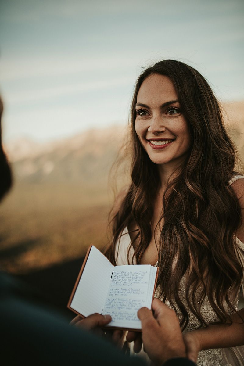 A photo showing a bride with long dark hair outdoors holding her grooms hand as he reads his vows to her.