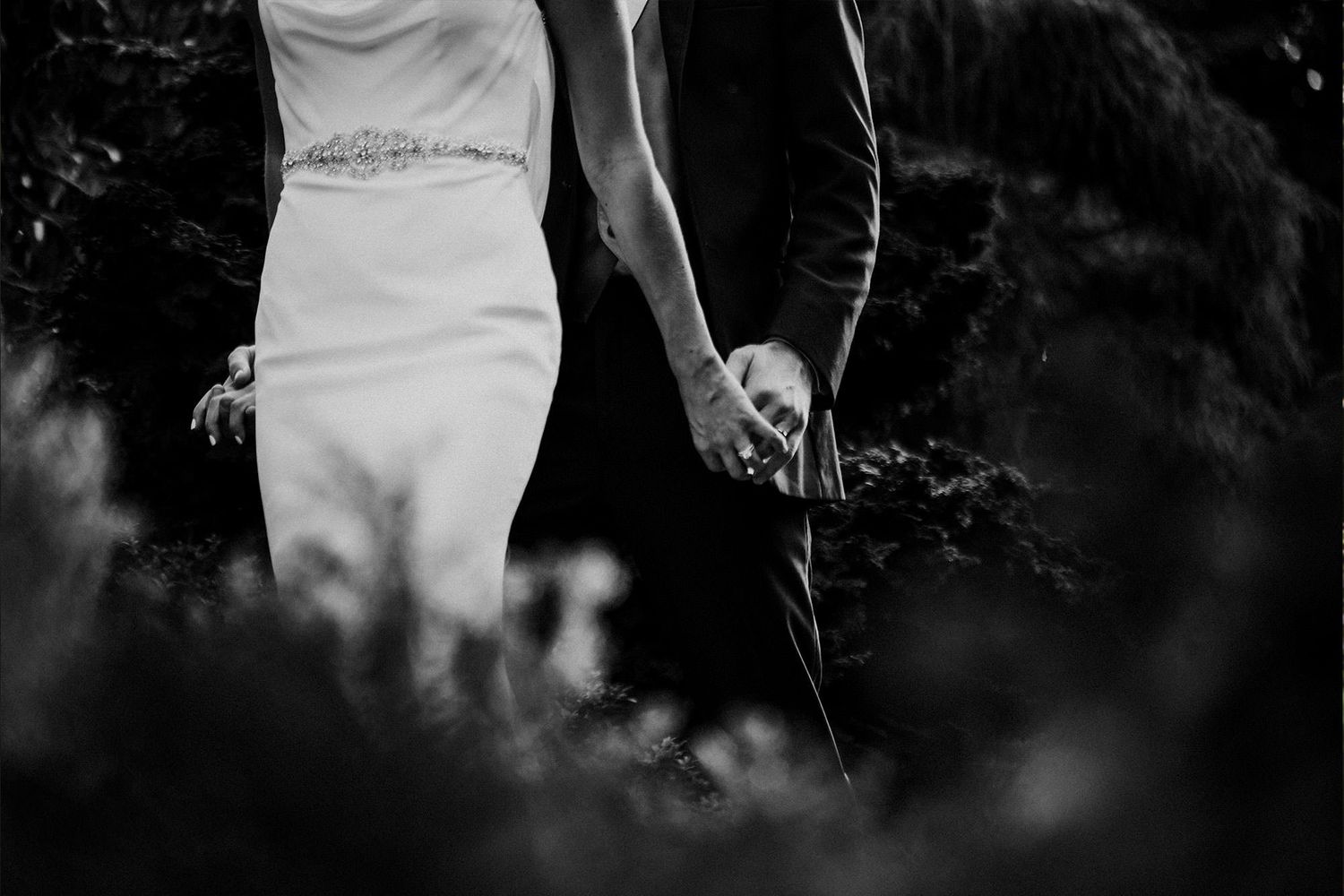 A black and white photo of a bride in white dress leading her groom through a dark forest setting.