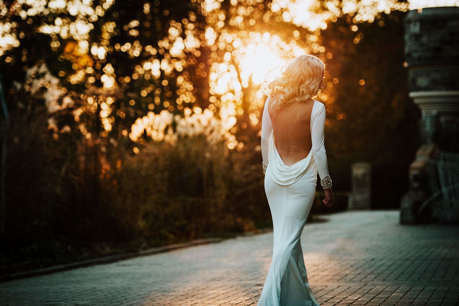 A bride in a white dress walks away from the camera in a garden setting during golden hour sunset with glowing backlight.