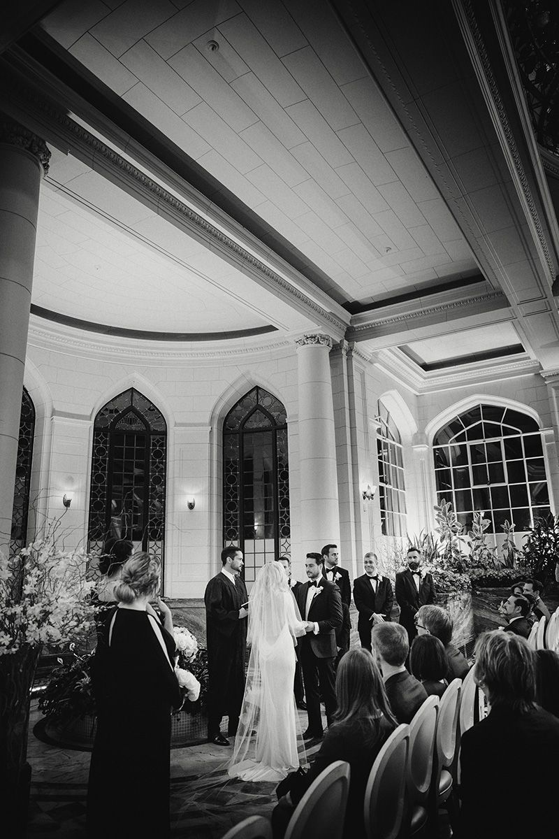 Black and white photo showing a wedding ceremony procession in an elegant room with arched windows and white columns.