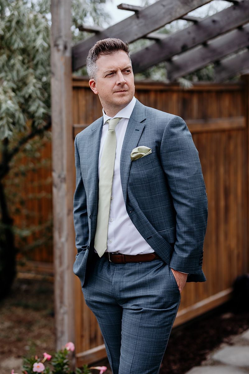 Stylish groom in tailored blue-grey suit and cream tie poses in garden setting with wooden pergola backdrop.