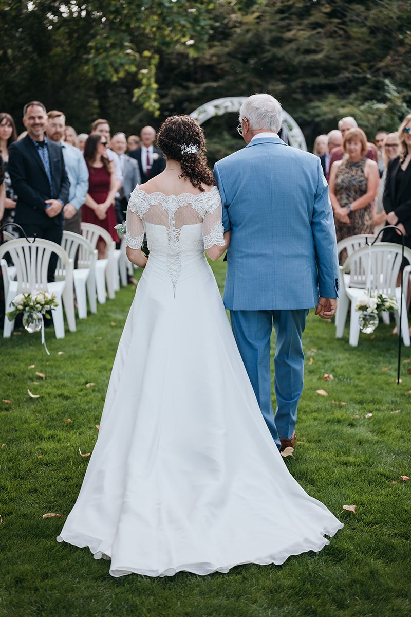 A bride in a lace-back wedding dress walks down the aisle with her father in a blue suit at an outdoor garden ceremony.