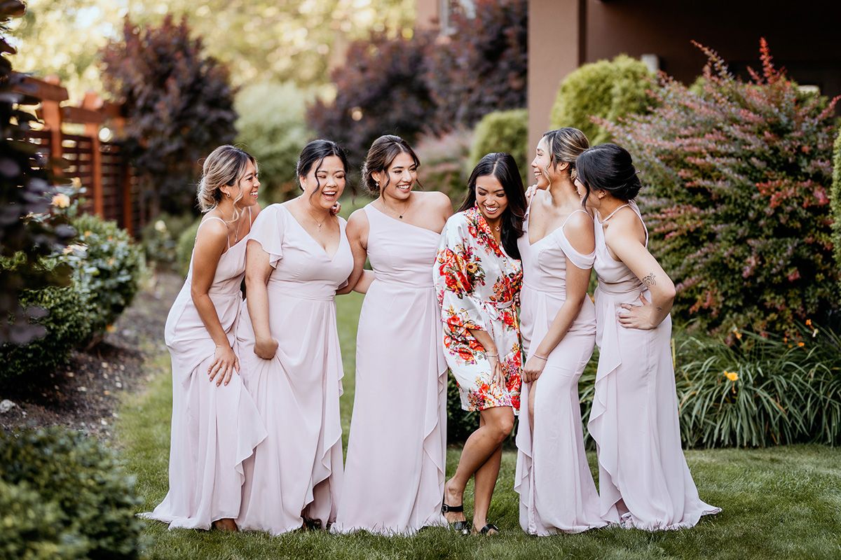 A bridal party in pink dresses along with a bride in her robe laughs together while standing in a garden setting.