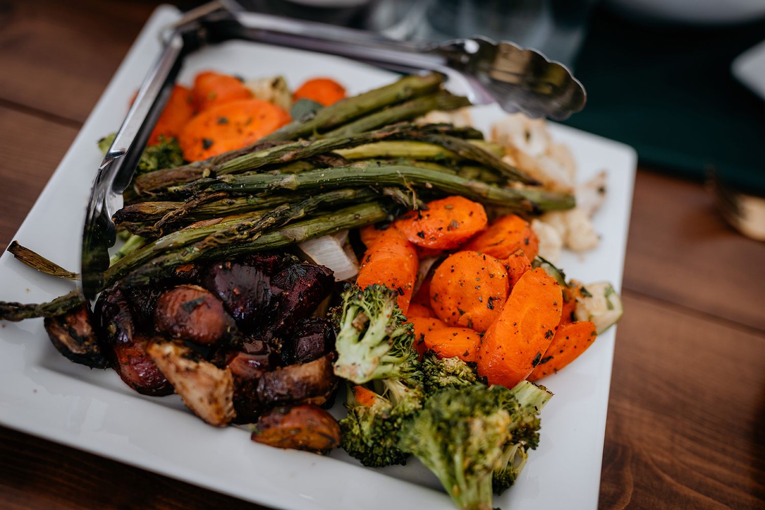 Roasted vegetables including carrots, asparagus, broccoli, and mushrooms arranged on white plates on a wooden table.