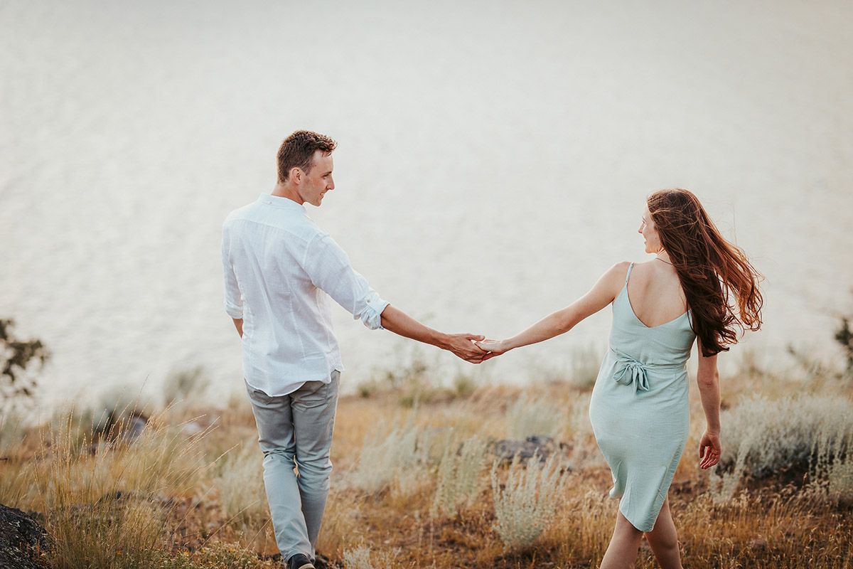 A couple holds hands while walking through a scenic field near a misty lake, the woman in a mint dress leading the way.