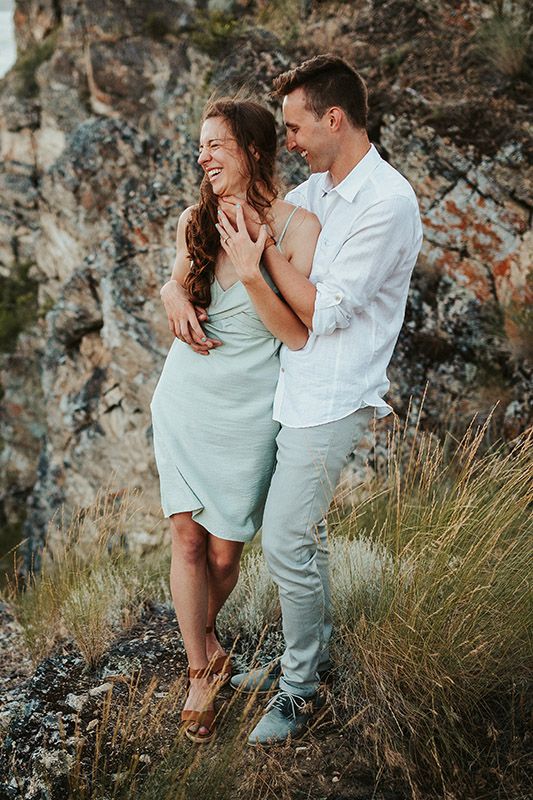 Happy couple embracing outdoors near rocky cliff, woman in mint dress laughing with man in white shirt and light pants.