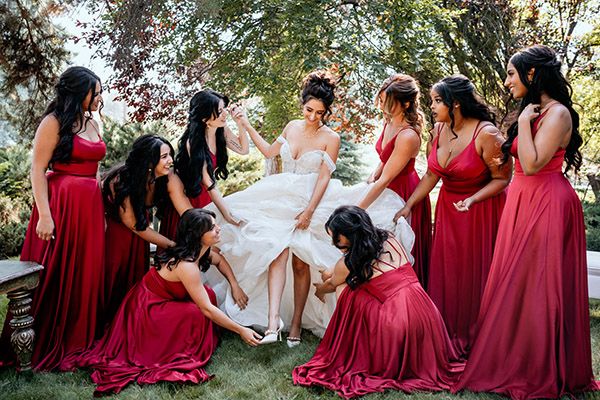 In an outdoor garden setting, a wedding party in burgundy dresses help a bride put on her shoes.