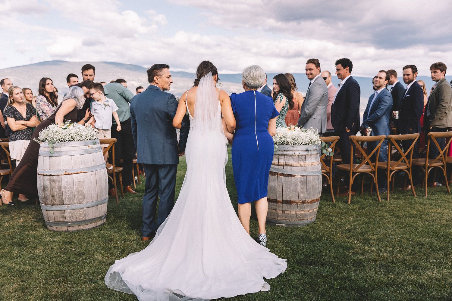 Wedding guests gather outdoors around rustic wine barrels during a vineyard celebration at sunset.