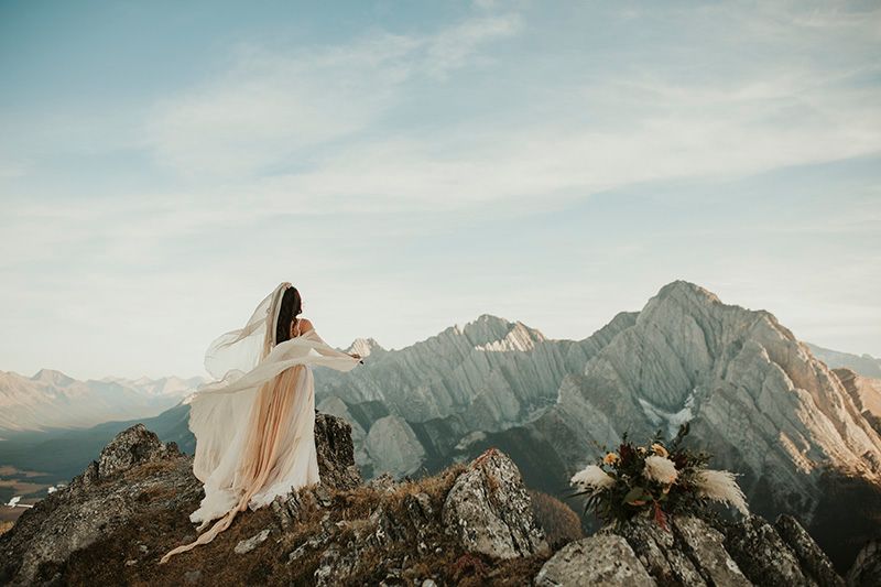 A bride in a wispy wedding dress stands atop rugged mountain peaks with a soft golden glow during sunset in a dramatic landscape.