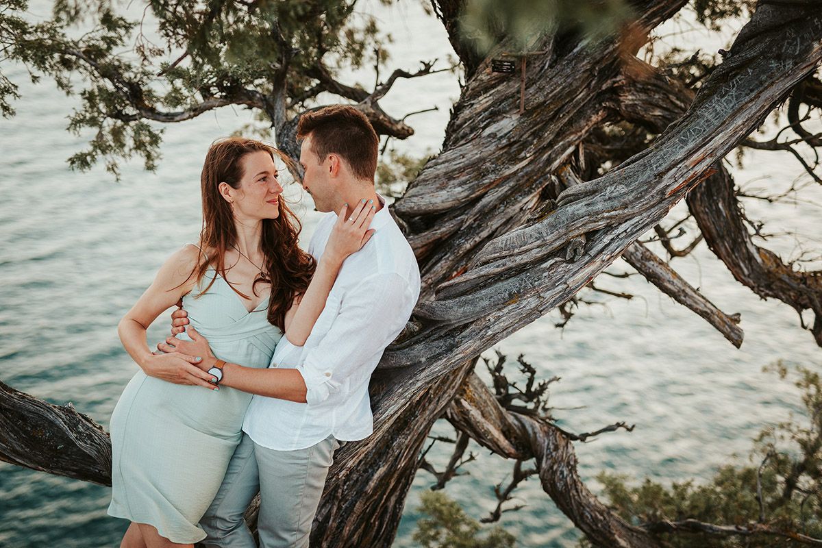 Romantic couple embracing near a gnarled tree by the sea, sharing an intimate moment outdoors.
