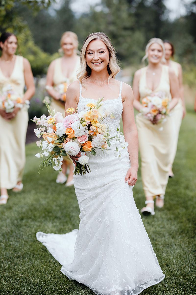 Wedding party in yellow bridesmaid dresses poses outdoors with beautiful floral bouquets featuring peach and white blooms.